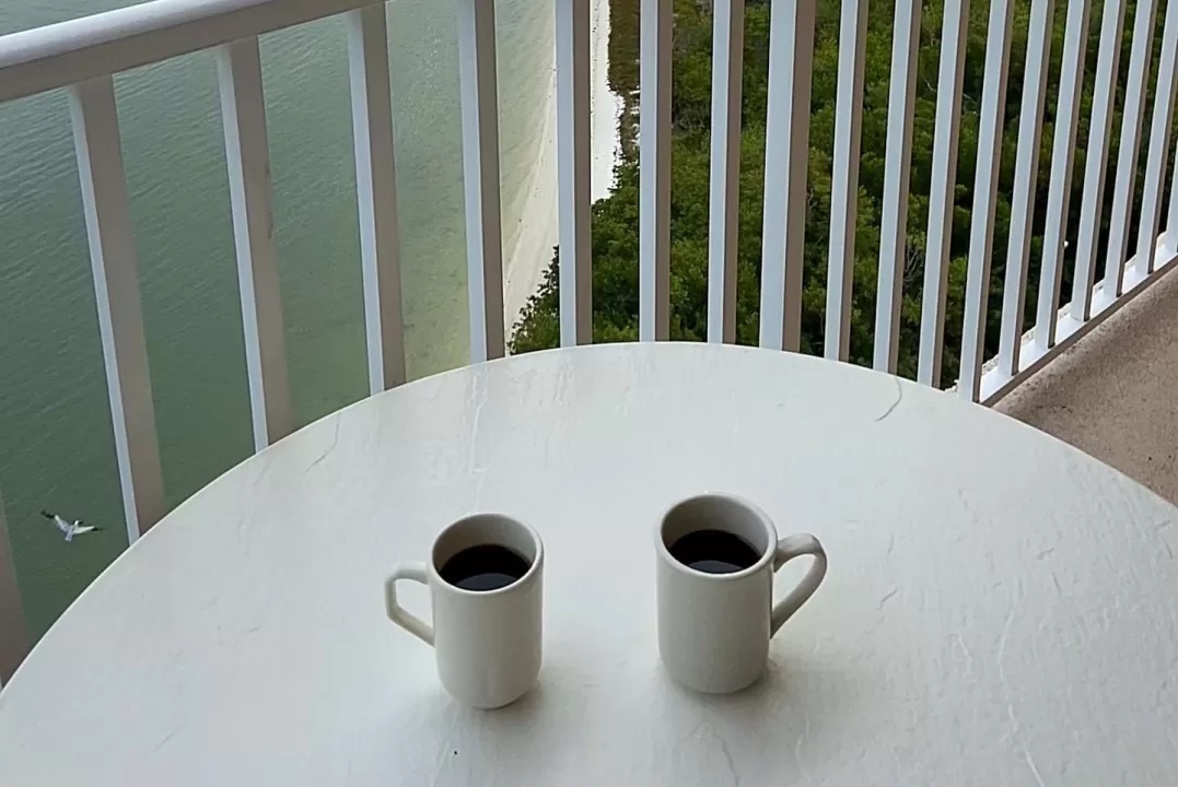 Zwei Kaffeetassen sitzen auf einem Tisch im Freien auf einem Balkon mit Blick auf Wasser und Sonnenuntergang
