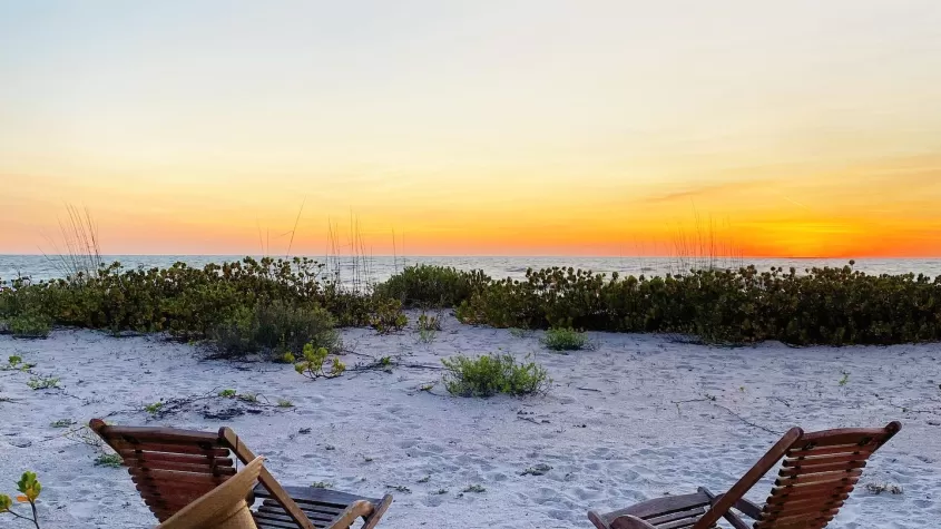 dos sillas de playa de madera vacías frente a la arena de la playa y la puesta de sol al fondo