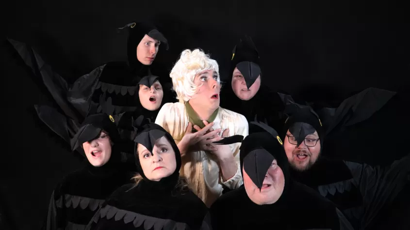 Una imagen de un grupo de actores vestidos como cuervos rodeando a un actor vestido como Melonie Daniels con cara de miedo.