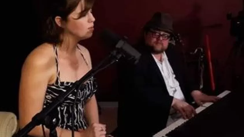 woman sitting in a chair in a strapless gown singing into a mic with a man in a suit and top hat playing piano
