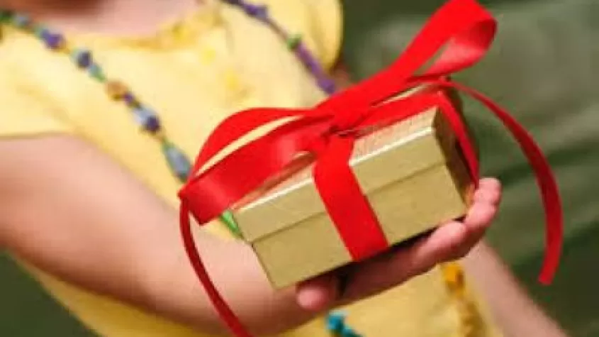 a child in a yellow shirt holding out a wrapped gift in gold paper and a red bow
