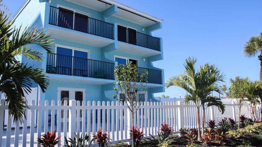 Bay inn  blue building exterior with landscaping, palm trees, white fence, and blue skies