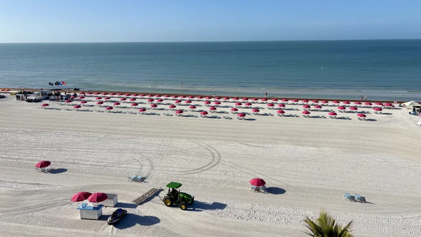 rooftop view overlooking the beach, pink umbrellas and beach chairs, palm trees, blue water, and blue skiies