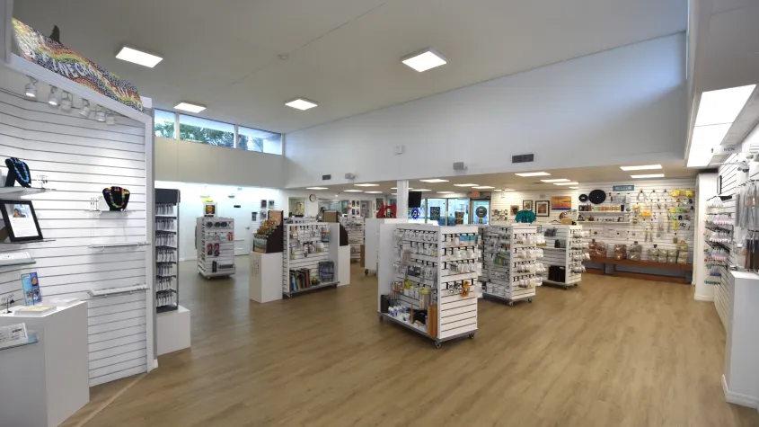 Spacious art supply store with white merchandise displays, wood-look flooring, and a gray bench positioned near the center. Art supplies and colorful merchandise fill the surrounding shelves.