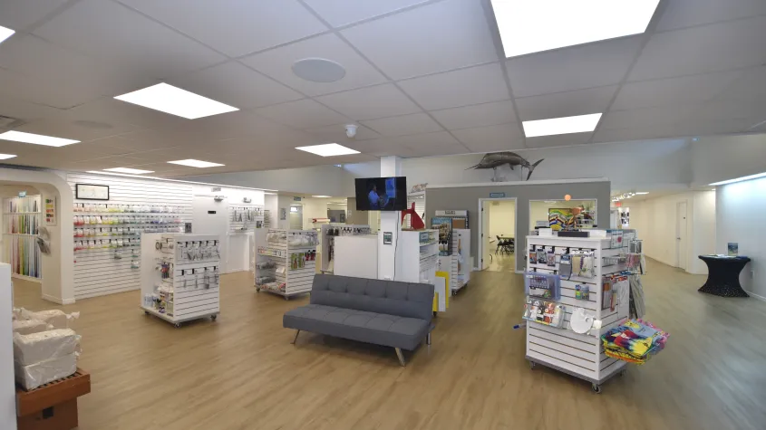 Wide view of a bright art supply store featuring rows of white merchandise displays stocked with art materials. Artwork and additional products line the walls, with natural light entering through high windows.