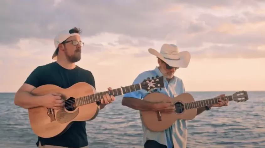 2 men walk with their guitars as they perform a song on the beach
