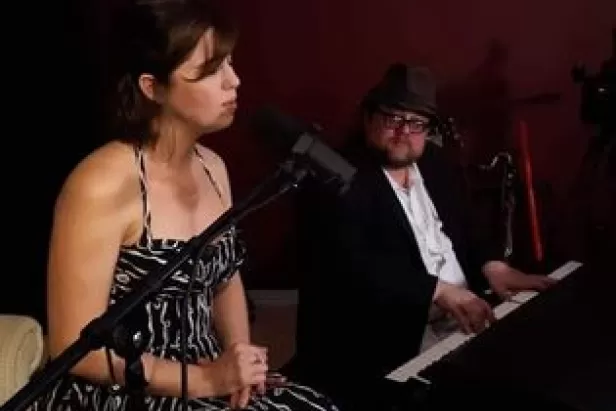 woman sitting in a chair in a strapless gown singing into a mic with a man in a suit and top hat playing piano
