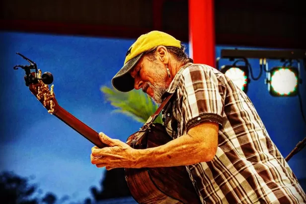 man in a baseball cap and brown striped short sleeve playing an acoustic guitar while standing in front of a night sky
