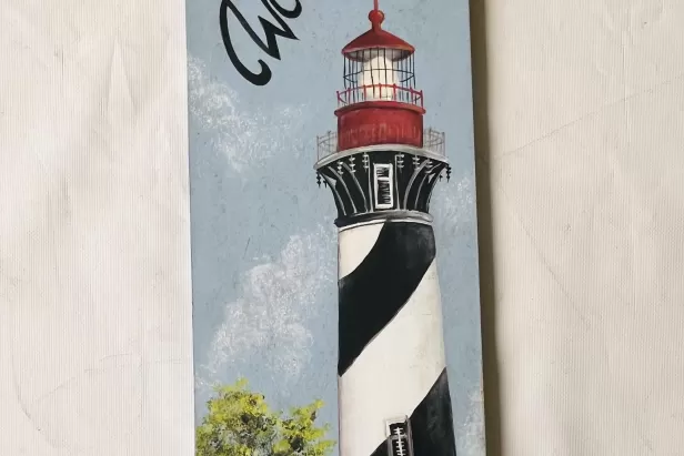 Narrow canvas with a black and white striped light house with a red top and green bushes at the base
