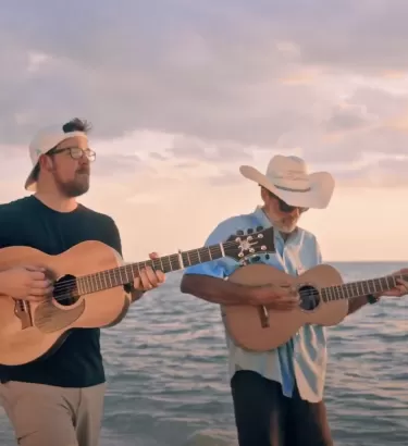2 men walk with their guitars as they perform a song on the beach
