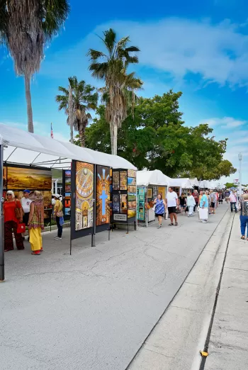 Visitors stand at an outdoor art fair booth examining framed watercolor paintings of birds and coastal wildlife displayed on a black gallery wall, with tents and other attendees visible in the background.