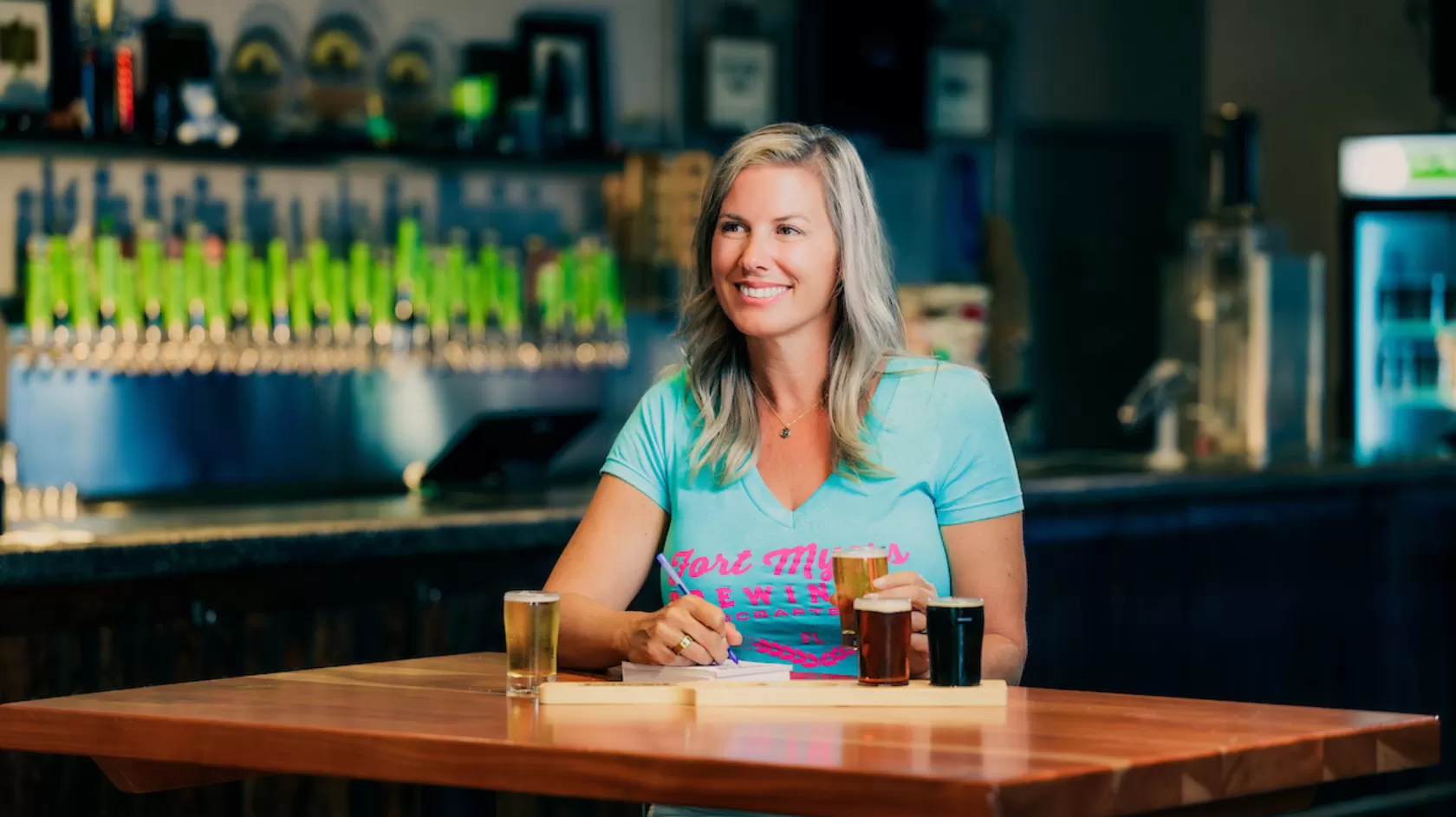 A woman sits at a high top table at Fort Myers Brewing Co with a flight of beer