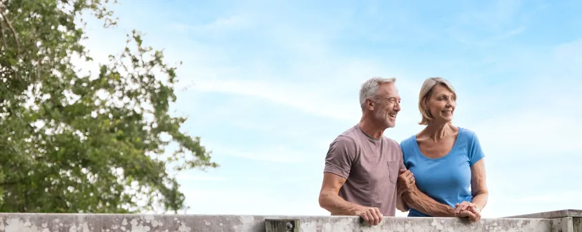 A smiling couple pauses on a wooden boardwalk surrounded by leafy trees and blue sky.