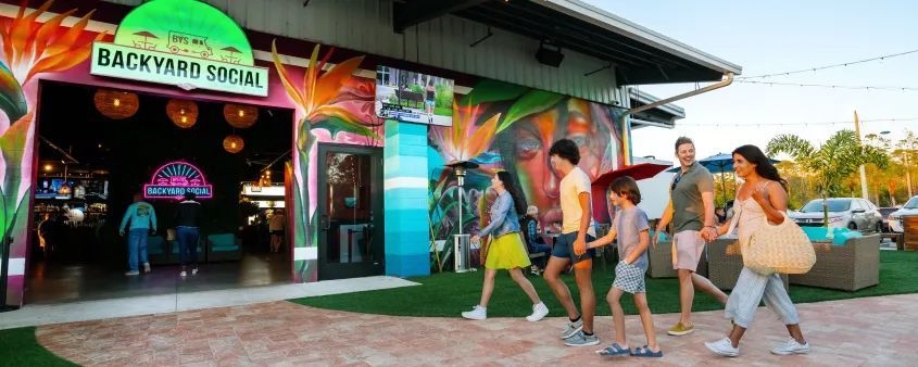 A family walks into Backyard Social in Fort Myers beneath colorful murals and outdoor string lights.