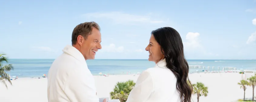 A couple in robes shares coffee on a beachfront hotel balcony.