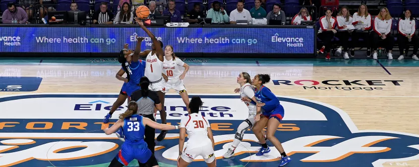 Women's college basketball players jump for a tip-off on a court branded with Fort Myers tournament logos.