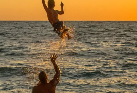 A dad throws his son up in the air over the water as the sun sets on the beach.