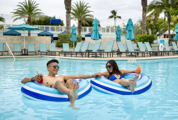 Man and woman floating in a resort pool on striped inner tubes, holding colorful drinks and smiling at each other.