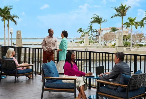 A group of meeting attendees talks on an outdoor waterfront terrace overlooking a marina lined with palm trees.