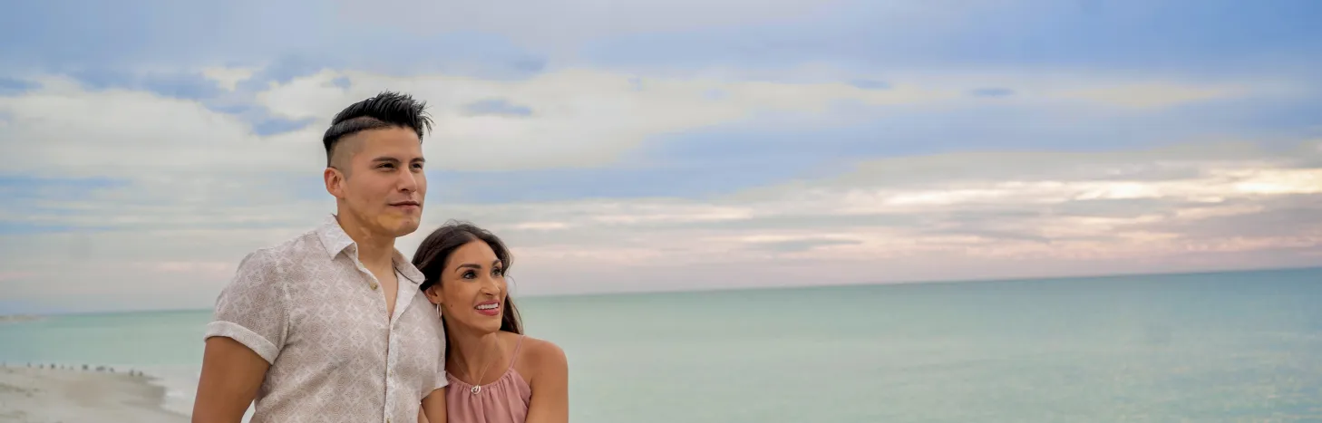 Couple standing hand in hand on a sandy beach at sunset, looking out over calm turquoise water under a cloudy sky.