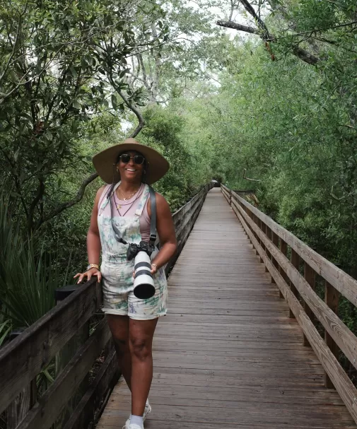 Smiling woman in a straw hat with a camera standing on a wooden boardwalk in the forest.