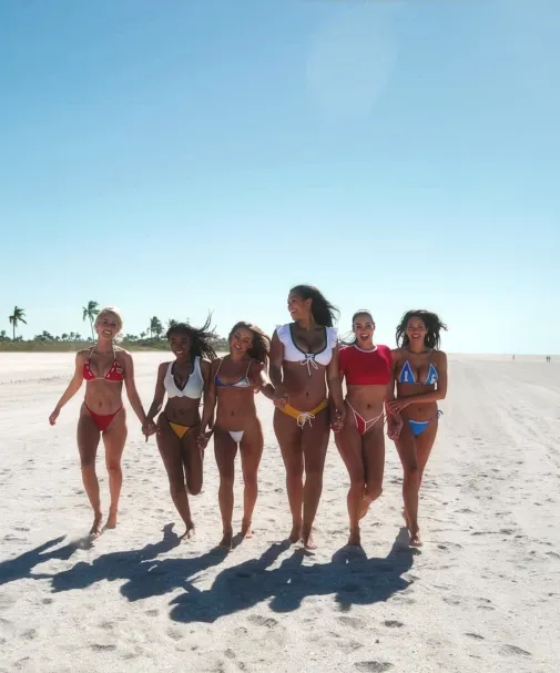 Six women in colorful bikinis walk hand in hand along a wide sandy beach under a clear blue sky.