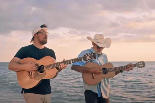 2 men walk with their guitars as they perform a song on the beach