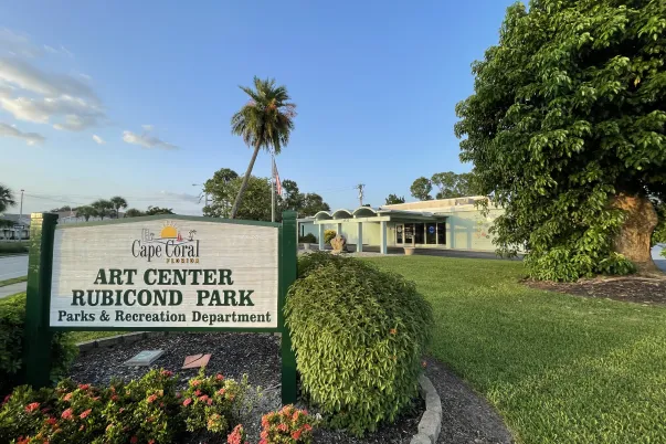 Photo of the Cape Coral Art Center at Rubicond Park on a sunny day. A large entrance sign reading “Cape Coral Art Center, Rubicond Park, Parks & Recreation Department” stands among colorful landscaping, with the Art Center building 2 in the background, green lawn, palm trees, and a large shade tree visible in the background.