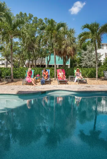 Four women lounging on poolside chairs beneath palm trees on a sunny day, chatting and reading beside a turquoise swimming pool.