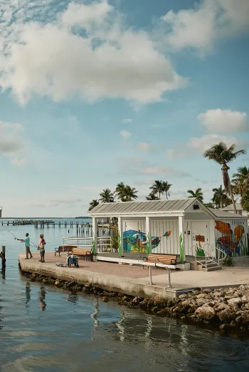Two people fish from the waterfront at Bat House Park in Matlacha beside a small mural-covered building.